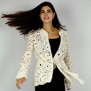 May include: Ivory crochet jacket with long sleeves and a tie closure. The jacket features a detailed openwork design with circular and floral patterns. The model is wearing a black top underneath.