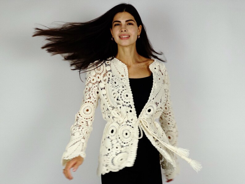 May include: Ivory crochet jacket with long sleeves and a tie closure. The jacket features a detailed openwork design with circular and floral patterns. The model is wearing a black top underneath.