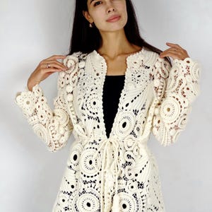 May include: Cream-colored, open-front crochet jacket with intricate circular and floral patterns. The jacket features long sleeves, a tie waist, and a delicate tassel detail. The garment is made of a soft, open-weave material, perfect for layering.