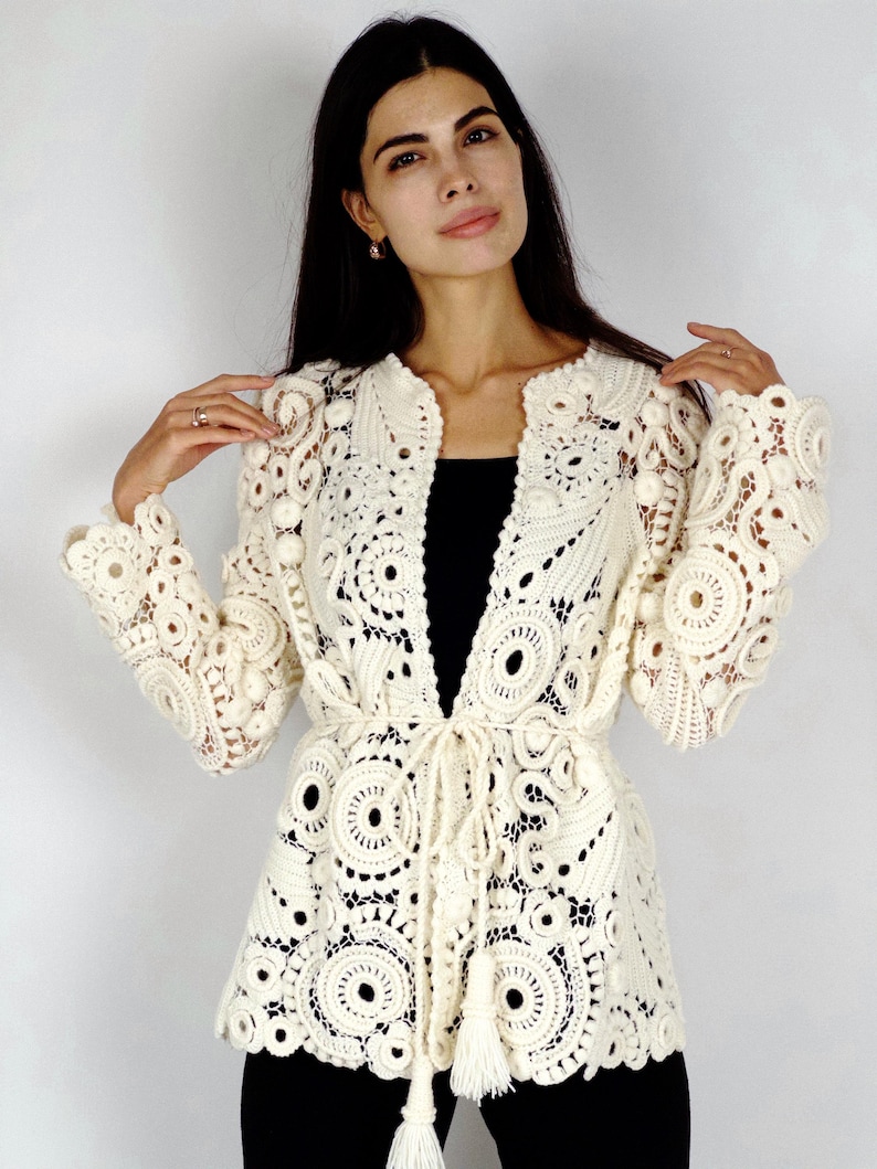 May include: Cream-colored, open-front crochet jacket with intricate circular and floral patterns. The jacket features long sleeves, a tie waist, and a delicate tassel detail. The garment is made of a soft, open-weave material, perfect for layering.