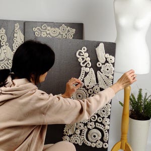 May include: A person working on a white crochet design on a dark gray background. The design features floral and geometric patterns. A dress form and a small potted plant are in the background.