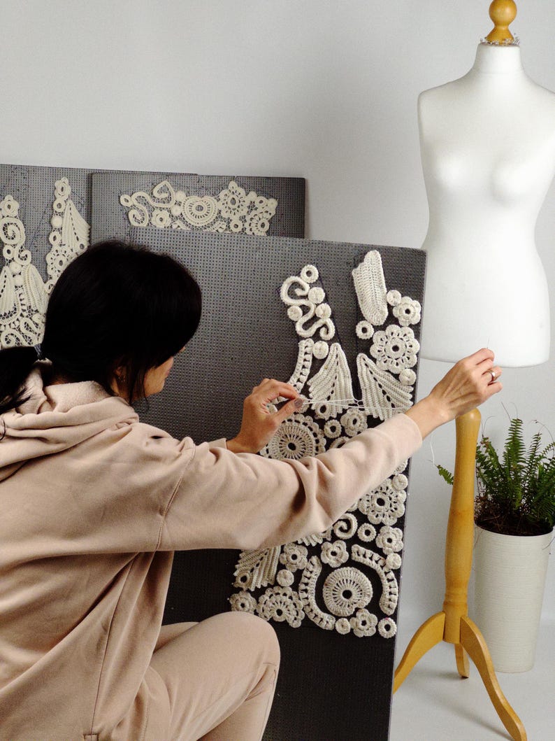 May include: A person working on a white crochet design on a dark gray background. The design features floral and geometric patterns. A dress form and a small potted plant are in the background.