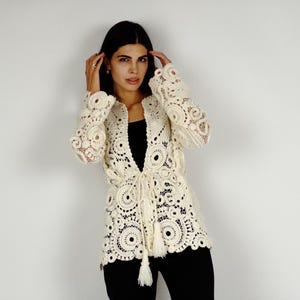 May include: Cream-colored, open-front crochet jacket with intricate circular and floral patterns. The jacket features long sleeves and a tie closure at the waist with tassels. The model is wearing black pants and a black top. The jacket is a unique piece of clothing.