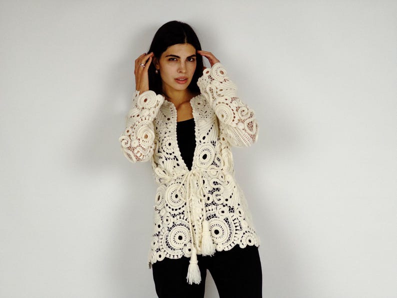 May include: Cream-colored, open-front crochet jacket with intricate circular and floral patterns. The jacket features long sleeves and a tie closure at the waist with tassels. The model is wearing black pants and a black top. The jacket is a unique piece of clothing.