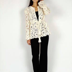 May include: Cream-colored, open-front crochet jacket with intricate circular patterns and long sleeves. The jacket features a tie closure at the waist with a tassel detail. The model is wearing black pants. This jacket is a stylish addition to any wardrobe.