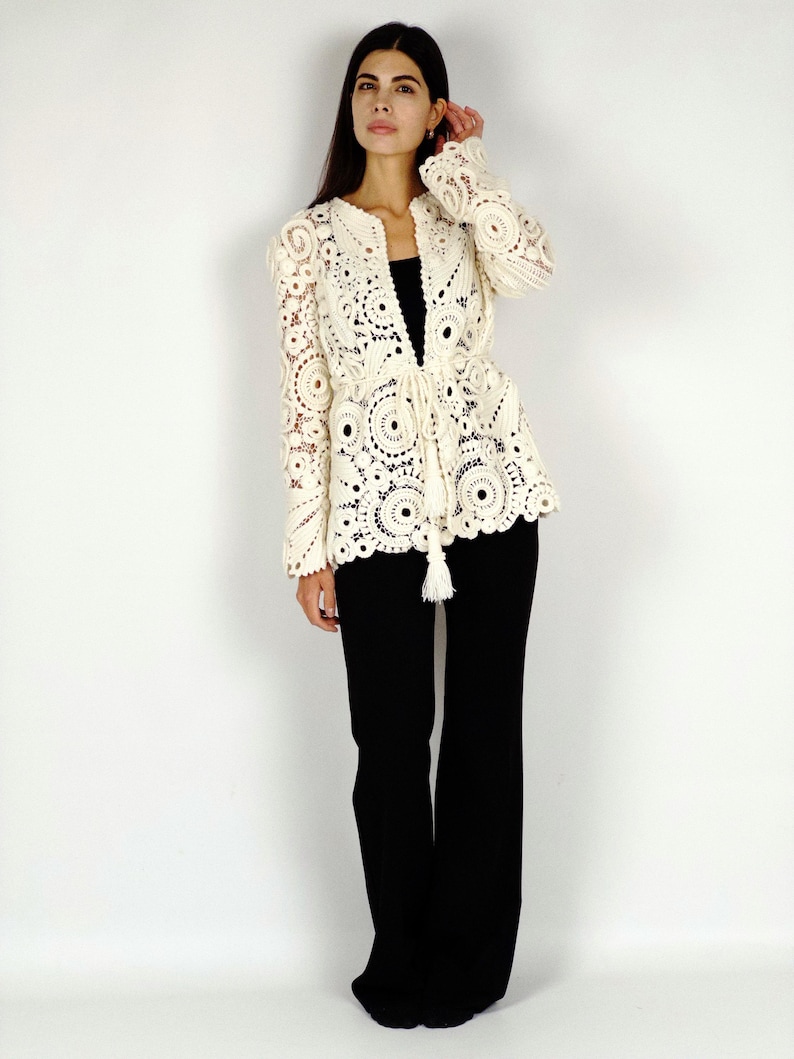 May include: Cream-colored, open-front crochet jacket with intricate circular patterns and long sleeves. The jacket features a tie closure at the waist with a tassel detail. The model is wearing black pants. This jacket is a stylish addition to any wardrobe.