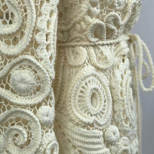 May include: Close-up of an off-white, handmade crochet garment. The intricate design features repeating circular and spiral patterns, with a textured, open weave. A belt cinches the waist, and the garment appears to be a dress or a long cardigan.