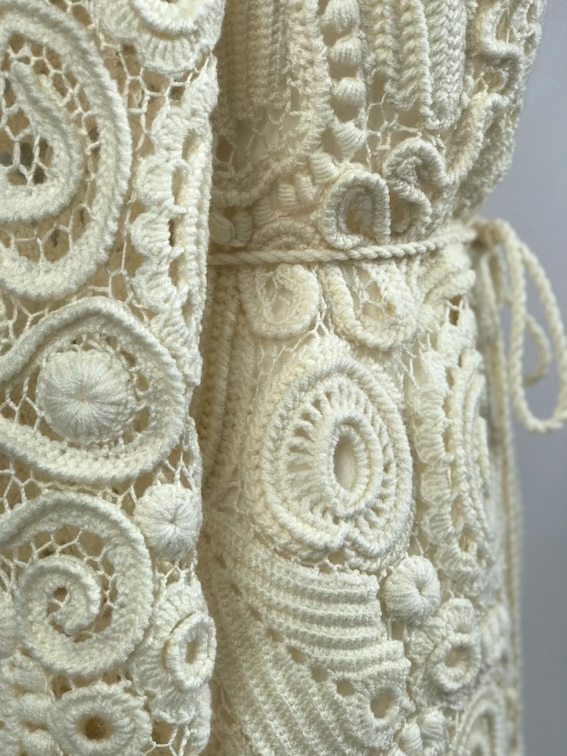 May include: Close-up of an off-white, handmade crochet garment. The intricate design features repeating circular and spiral patterns, with a textured, open weave. A belt cinches the waist, and the garment appears to be a dress or a long cardigan.