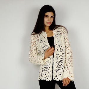 May include: Cream-colored, open-front crochet jacket with intricate circular and floral patterns. The jacket features long sleeves and a collarless design. The garment is worn over a black top and black pants.