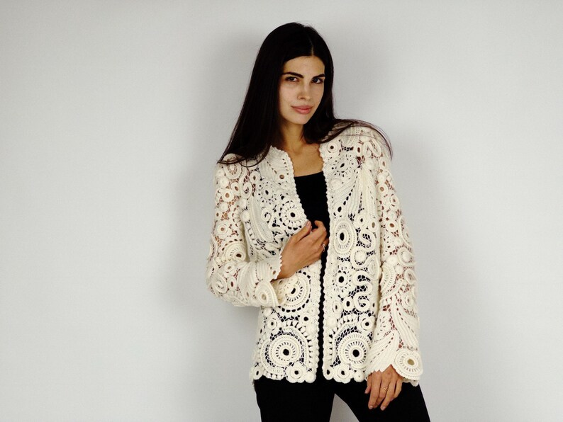 May include: Cream-colored, open-front crochet jacket with intricate circular and floral patterns. The jacket features long sleeves and a collarless design. The garment is worn over a black top and black pants.