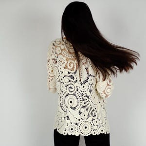 May include: Cream-colored crochet top with intricate circular and floral patterns. The top has long sleeves and a scalloped hem. The person is wearing black pants. The background is a plain white wall.