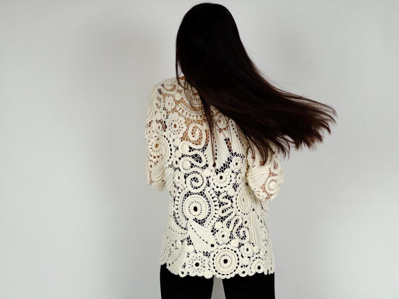 May include: Cream-colored crochet top with intricate circular and floral patterns. The top has long sleeves and a scalloped hem. The person is wearing black pants. The background is a plain white wall.