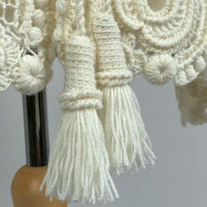 May include: Close-up of a cream-colored, handmade crochet item. The piece features intricate patterns, including circular designs and textured bobbles. Two tassels hang down, adding a decorative element to the textile.