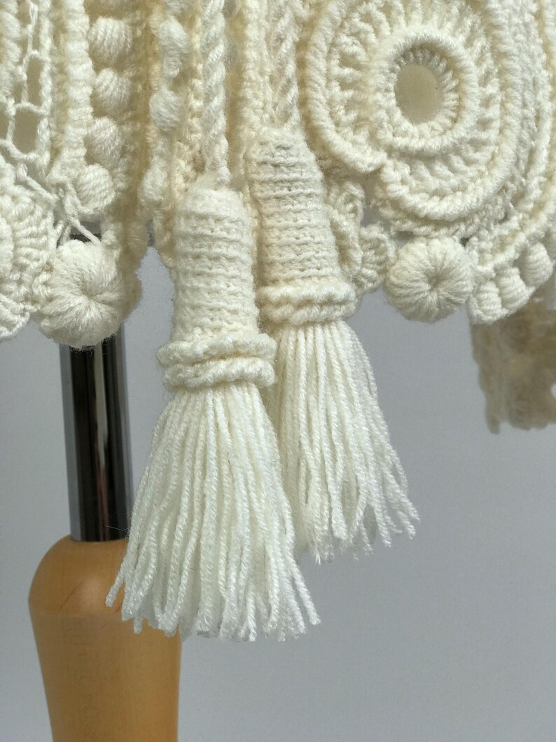 May include: Close-up of a cream-colored, handmade crochet item. The piece features intricate patterns, including circular designs and textured bobbles. Two tassels hang down, adding a decorative element to the textile.