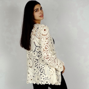 May include: Cream-colored, open-weave crochet jacket with long sleeves. The jacket features a detailed floral and circular pattern. The garment is worn over a black top and black pants. The jacket's intricate design is visible.