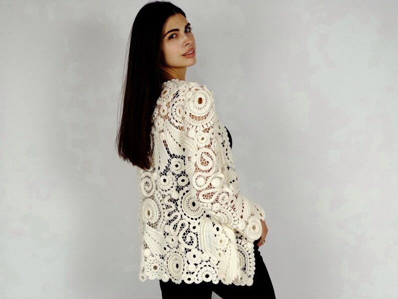 May include: Cream-colored, open-weave crochet jacket with long sleeves. The jacket features a detailed floral and circular pattern. The garment is worn over a black top and black pants. The jacket's intricate design is visible.