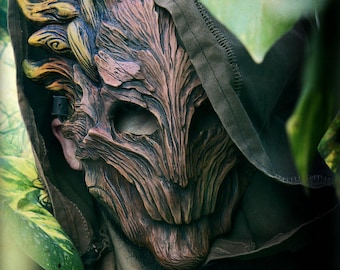 May include: A brown and tan textured mask with a wood-like grain pattern, featuring deep eye sockets and a jagged mouth area. The mask is worn under a dark brown hooded garment. The text CREADORES A SUELDO CREATORS FOR HIRE is visible at the bottom.