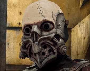 Könnte beinhalten: Eine Vollkopfmaske mit einem verwitterten, cremefarbenen, schädelartigen Oberteil mit sichtbaren Rissen und metallischen mechanischen Komponenten um Augen und Kiefer. Der untere Gesichtsbereich hat ein dunkles, texturiertes Gitter mit kreisförmigen Perforationen.