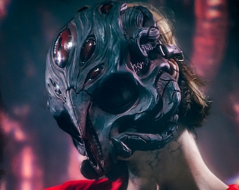 May include: A person wearing a red shirt and a dark gray mask with multiple eyes. The mask has a textured, organic design and appears to be made of a hard material. The text "CREATURES OF THE SUELO" is visible on the shirt.