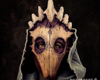 May include: A handcrafted, beige and purple mask with a bird-like skull design. The mask features a crown-like structure and is worn with a blue hooded garment. The eyes of the wearer are visible through the eye sockets. The mask has a weathered, aged appearance.