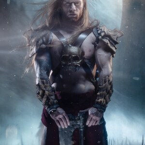 May include: A long-haired warrior with a beard wearing dark leather chest straps featuring a central skull ornament and textured bone-like shoulder and forearm armor. The figure holds a metallic battle axe and wears a dark red cloth garment.