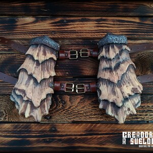 May include: A pair of arm guards featuring layered, scale-like plates in shades of tan and dark brown with a textured, hammered metal-style top edge. The guards are secured with brown leather straps and brass-toned metal buckles.