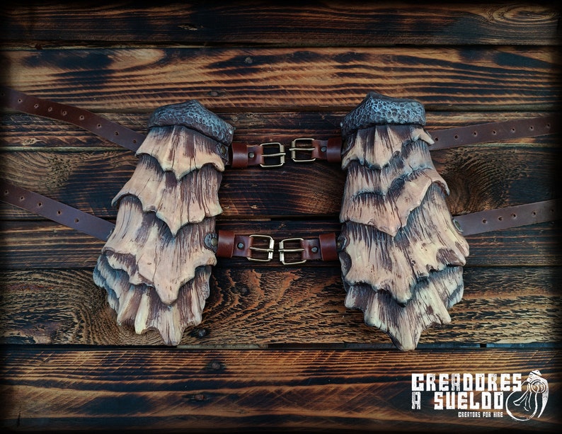 May include: A pair of arm guards featuring layered, scale-like plates in shades of tan and dark brown with a textured, hammered metal-style top edge. The guards are secured with brown leather straps and brass-toned metal buckles.