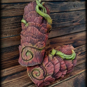 May include: Two sculpted arm guards featuring a layered, bark-like texture in shades of reddish-brown and deep pink. Each piece displays a prominent spiral carving and is accented with bright green, vine-like tendrils wrapping around the surface.