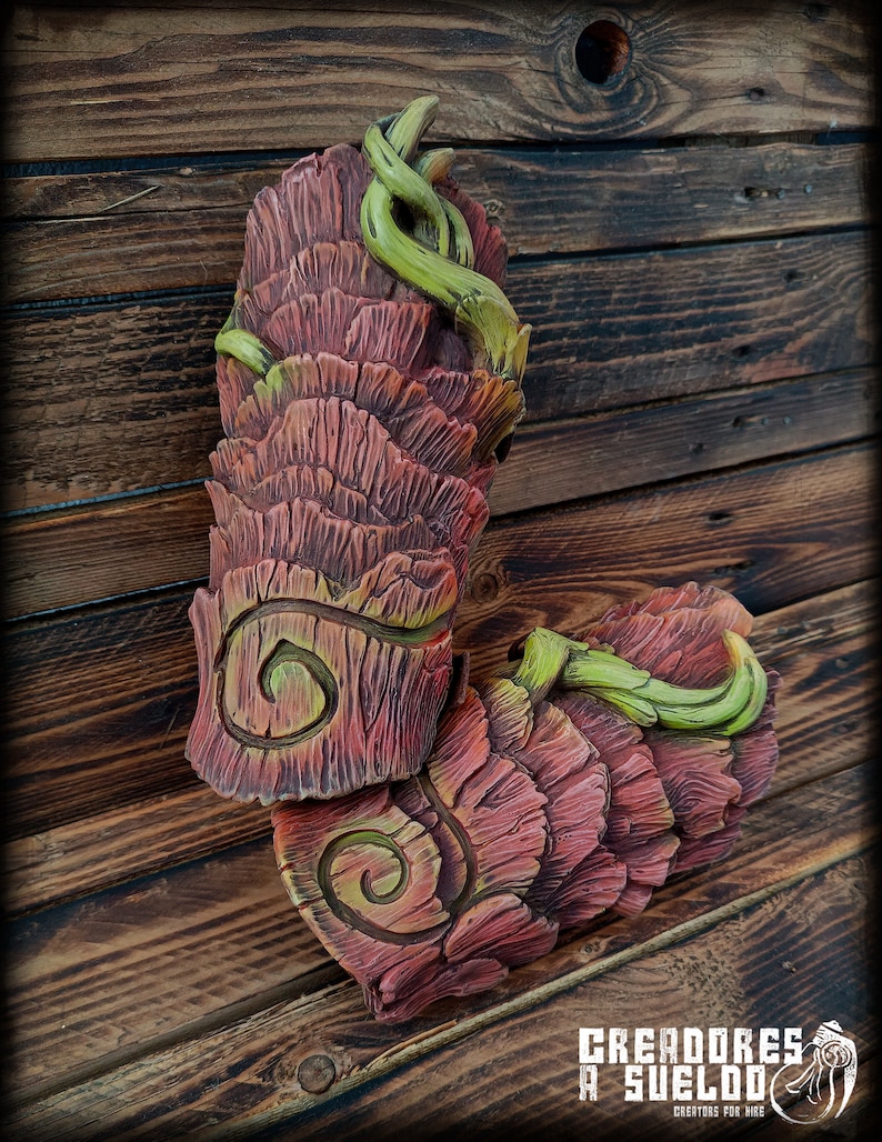 May include: Two sculpted arm guards featuring a layered, bark-like texture in shades of reddish-brown and deep pink. Each piece displays a prominent spiral carving and is accented with bright green, vine-like tendrils wrapping around the surface.