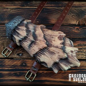 May include: A forearm guard featuring a layered, bark-like texture in shades of tan and dark brown, secured with two brown leather straps and brass-toned metal buckles. The text CREADORES A SUELDO CREATORS FOR HIRE is printed in the bottom corner.