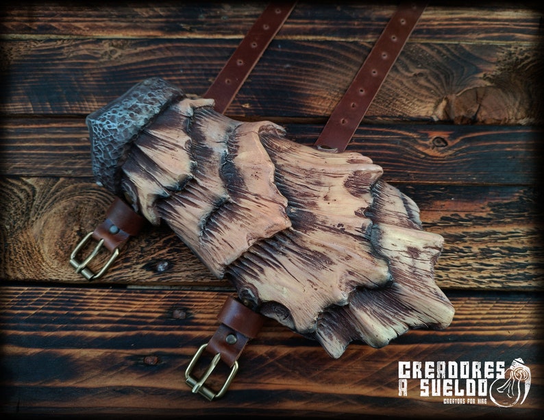 May include: A forearm guard featuring a layered, bark-like texture in shades of tan and dark brown, secured with two brown leather straps and brass-toned metal buckles. The text CREADORES A SUELDO CREATORS FOR HIRE is printed in the bottom corner.
