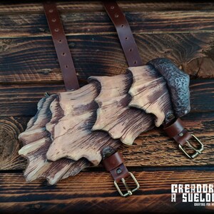 May include: A brown and tan layered arm guard featuring a textured, wood-like finish with a dark, hammered-effect top section. The piece includes two brown leather straps with brass-toned metal buckles for secure fastening to the arm.
