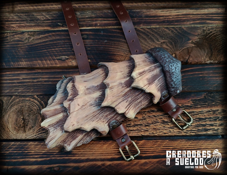 May include: A brown and tan layered arm guard featuring a textured, wood-like finish with a dark, hammered-effect top section. The piece includes two brown leather straps with brass-toned metal buckles for secure fastening to the arm.