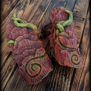 May include: A pair of arm guards featuring a textured, reddish-brown bark-like surface with carved spiral patterns and sculpted green vine accents. The guards include dark brown leather straps with metal buckles for securing the pieces to the arm.