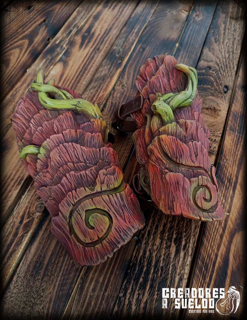 May include: A pair of arm guards featuring a textured, reddish-brown bark-like surface with carved spiral patterns and sculpted green vine accents. The guards include dark brown leather straps with metal buckles for securing the pieces to the arm.