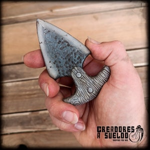 Puede incluir: Una daga de empuje con una hoja ancha y triangular de metal martillado gris oscuro y un mango texturizado gris claro con un patrón de veta de madera. El mango está asegurado con dos remaches visibles y tiene una forma curva y ergonómica.