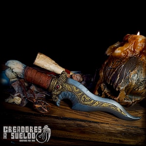 May include: A silver and gold dagger with a decorative handle and a red jewel. The dagger is lying on a wooden surface with a skull and a candle in the background.
