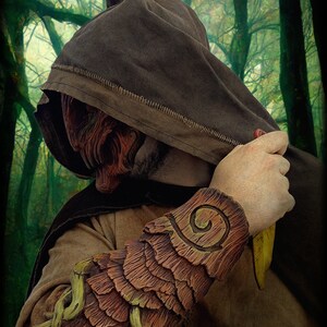 May include: A brown hooded cloak worn with a textured, bark-like forearm bracer featuring layered, wood-grain patterns and light green vine accents. The bracer displays a spiral carving on the upper section. The text CREADORES A SUELDO is visible.