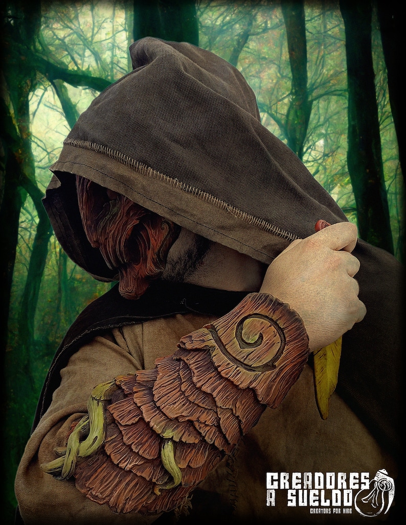 May include: A brown hooded cloak worn with a textured, bark-like forearm bracer featuring layered, wood-grain patterns and light green vine accents. The bracer displays a spiral carving on the upper section. The text CREADORES A SUELDO is visible.