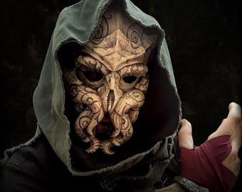 May include: A detailed, carved mask with a tentacled design, worn with a dark hooded cloak. The mask is tan with intricate patterns and dark eye sockets. The cloak is a muted green-gray color.