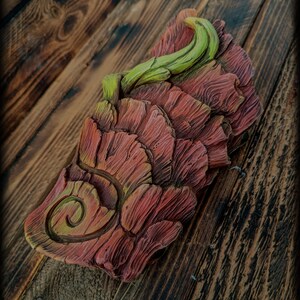 May include: A rectangular decorative object featuring a layered, bark-like texture in shades of reddish-brown and green. The surface displays a prominent spiral carving on the lower left and a curved, vine-like protrusion extending from the top.