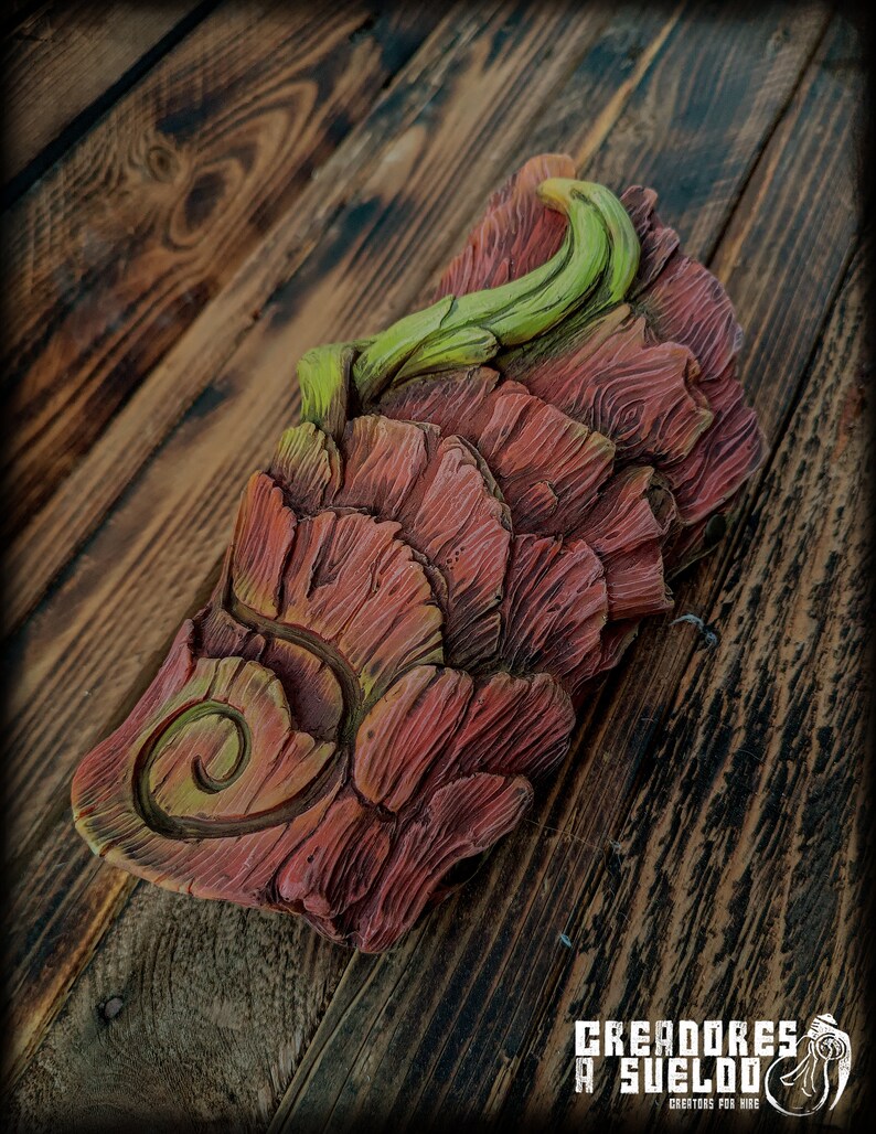 May include: A rectangular decorative object featuring a layered, bark-like texture in shades of reddish-brown and green. The surface displays a prominent spiral carving on the lower left and a curved, vine-like protrusion extending from the top.