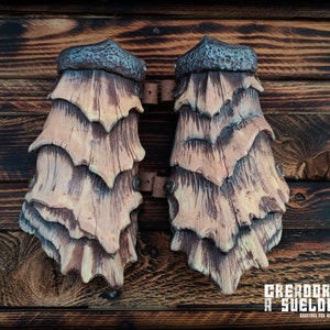 May include: A pair of tan and brown forearm guards featuring a layered, scale-like design with a textured, dark gray cuff at the top. The guards are connected by brown leather straps and metal rivets, displaying a wood-like grain pattern throughout.