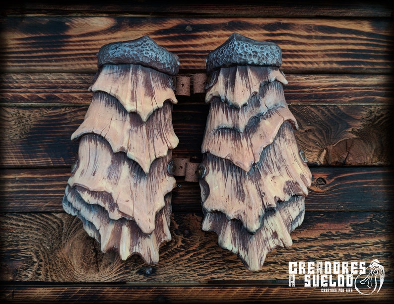 May include: A pair of tan and brown forearm guards featuring a layered, scale-like design with a textured, dark gray cuff at the top. The guards are connected by brown leather straps and metal rivets, displaying a wood-like grain pattern throughout.