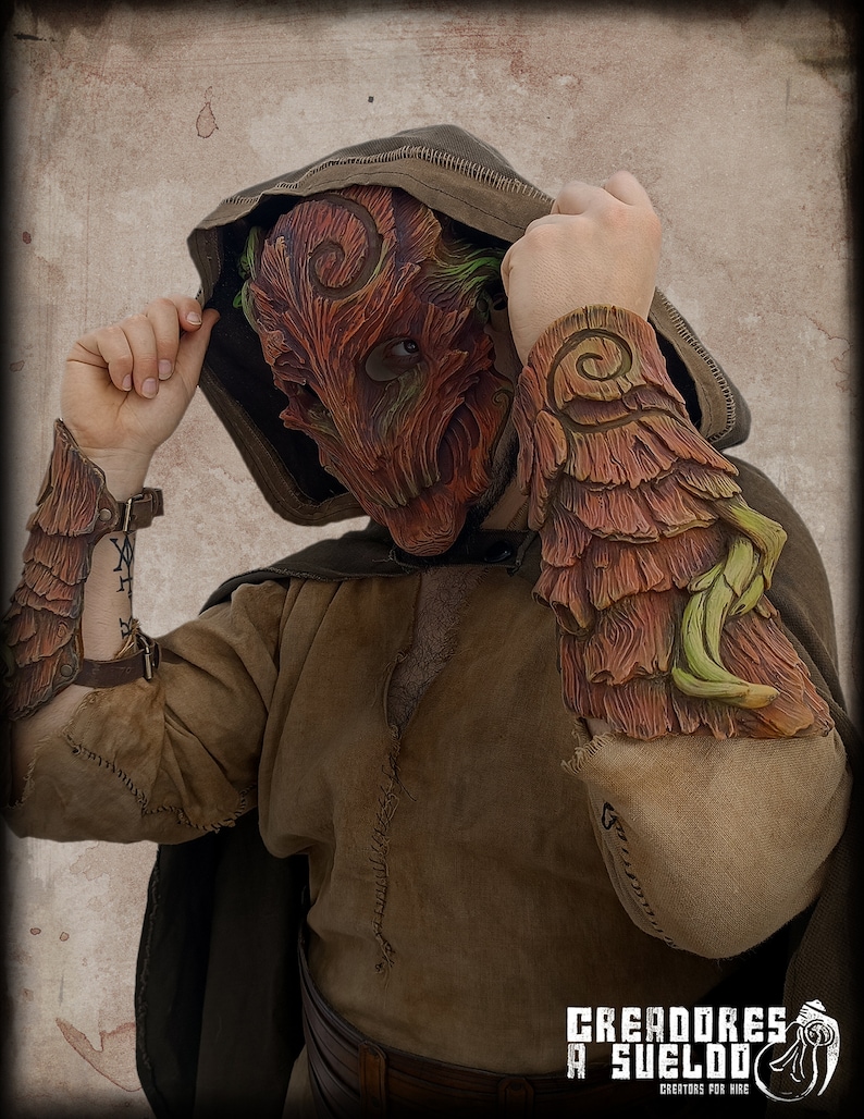 May include: A textured mask and matching forearm bracers featuring a carved wood-like pattern in shades of reddish-brown and green. The mask has spiral details and eye openings. The bracers are layered with leaf-like accents and brown straps.