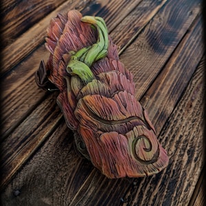 May include: A molded leather bracer featuring a textured bark-like surface in shades of reddish-brown and orange, accented with a raised, winding green vine element and a carved spiral detail. The piece includes a brown leather strap and buckle.