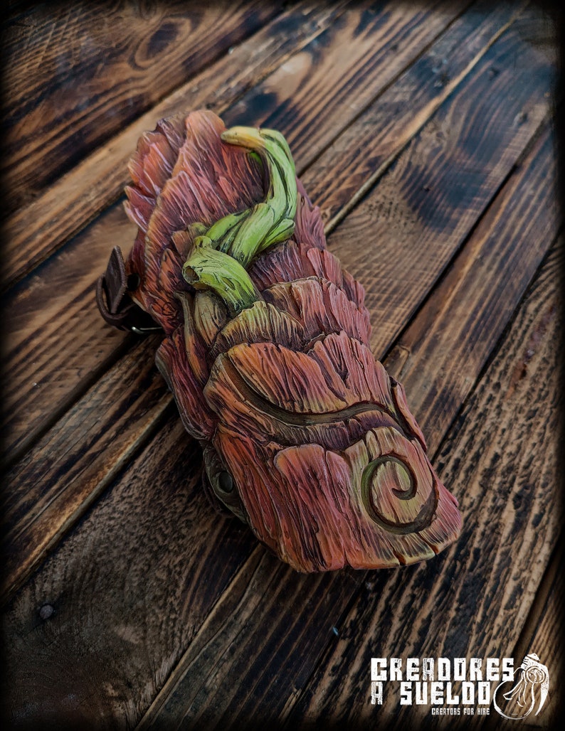 May include: A molded leather bracer featuring a textured bark-like surface in shades of reddish-brown and orange, accented with a raised, winding green vine element and a carved spiral detail. The piece includes a brown leather strap and buckle.