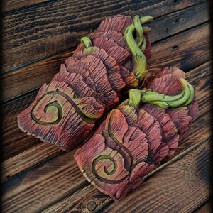 May include: A pair of forearm guards featuring a layered, bark-like texture in shades of reddish-brown. Each piece displays a prominent carved spiral design and is accented with sculpted, light green vine-like elements wrapping around the top edges.