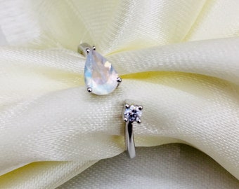 May include: A silver ring with a teardrop-shaped moonstone and a smaller round clear stone. The ring is on a white fabric background.