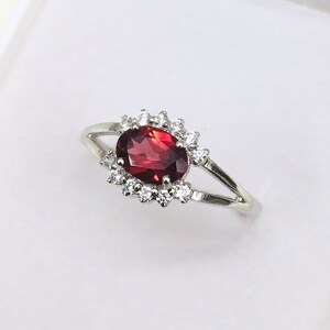Peut inclure: Une bague en argent ornée d'une grosse pierre rouge foncé de forme ovale, entourée d'un halo de petites pierres claires. Le corps de la bague se divise en deux brins délicats de chaque côté de la pierre centrale.